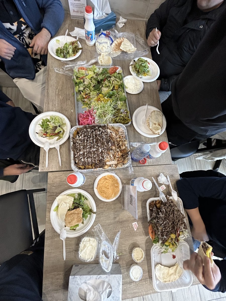 An overhead group spread of shawarma platters, salads, dips and drinks with several diners seated